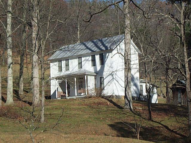 A two story white frame house with a porch in front and a metal roof sits in the middle of a grove of trees.