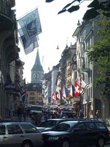 Zurich's main street is colored by many flags hanging from poles over various storefronts; it looks like flags of various Swiss cantons