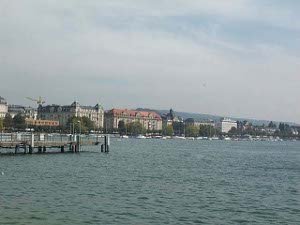 A pier is seen jutting out into the lake, and the eastern side of the Zurichsee south of the downtown area is lined with lakeside buildings; light clouds cover most of the sky