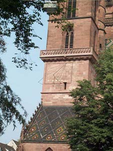 On the side of a reddish-tan stone church tower is an iron sundial