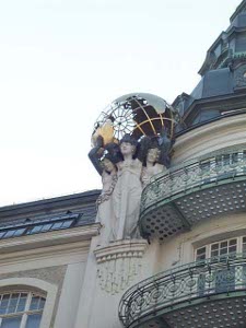 Near the roof of a building, next to a high metal balcony, is a statue of three women holding up a partly transparent globe in the art deco style