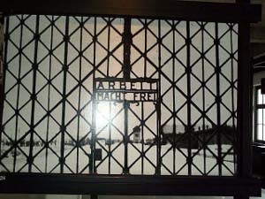 The sun shines in the sky behind the gate, with the words worked in wrought iron in the center of a lattice of vertical and diagonal iron bars, 'Arbeit Macht Frei'