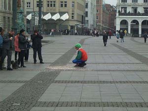 Wearing a red vest over a blue jacket with a bright green hat, the clown squats near a pile of confetti on the plaza