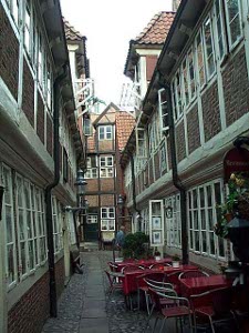A narrow passage with overhanging buildings on both sides, many windows, red tables set out for diners