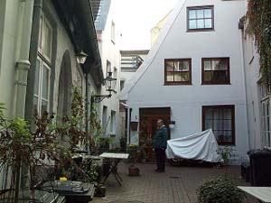 One building facing the courtyard is painted light green, with plants in pots; another is white with brown square windows and a steep roof; a motorcycle is covered with a sheet; a narrow alley passes to the left of the white building