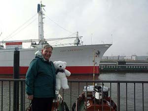 Bob stands holding Cuddles with a large white cargo ship with a red painted keel rides at anchor
