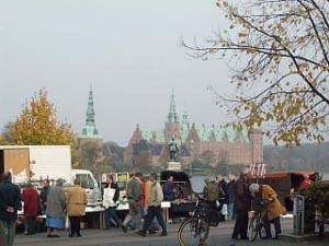 In the foreground a busy street with pedestrians, bicycles, trucks, market stalls; in the background, past the water, the reddish stone castle with copper green roofs