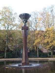 A 15 foor dark brown granite column supports a bronze cup overflowing with water tumbling into a large surrounding pool