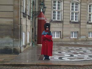 Dressed in a red overcoat with a black beaver hat, the guard has his arms folded for warmth and dryness
