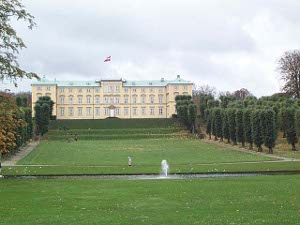 A three story tan stone castle with a light blue roof and the Danish flag flying high above the center, with terraces leading down to a long sloping lawn flanked by trees