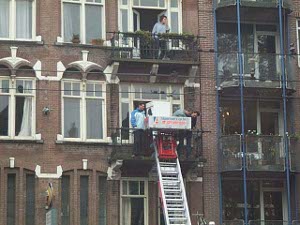 A long metal ladder has been attached to the second floor iron railing, and a motorized red jack supports the washer and climbs the ladder raising the washer to the upper floor where it enters the house through the window