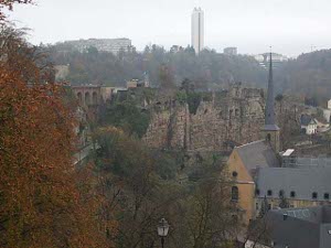 This panoramic view shows a steep descent from the heights, fortified walls, skyscrapers, and wooded hills, all in the city of Luxembourg