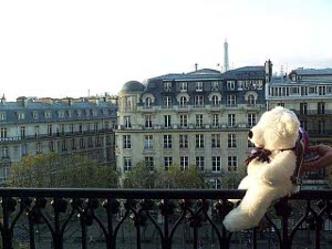 The teddy bear is balanced on the wrought iron railing high over the Champs Elysees; the morning sun shines brightly on the bear