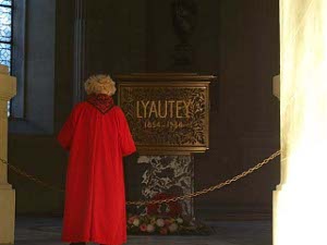 A blonde lady in a brilliant crimson full-length coat stands looking at the tomb of Marshal Hubert Lyautey at Les Invalides