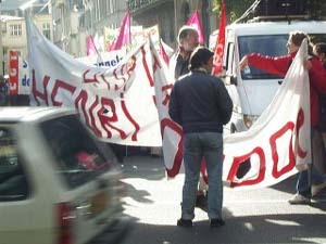 Demonstrators with large white banners bearing red letters maneuver into parade position past moving vehicles in preparation for a labor movement street demonstration