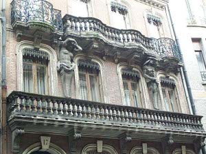 The second and third floors of this brick building have elaborate balconies and French windows.  Two caryatids are affixed to the walls supporting the third floor balcony.  They are of carved stone, gray with age, of different classical designs.