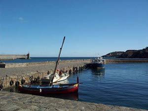A long stone pier juts out into the deep blue Mediterranean water; a few boats, including one Catalan fishing boat are tied up along the jetty.