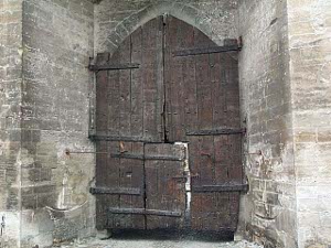 In a wide stone archway is a tall dark wooden door which hinges to both sides; inside this door is a small person size door at the center and bottom