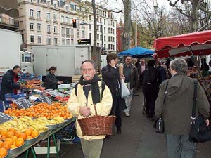 Oranges, bananas, tangerines are laid out.  A shopper in a yellow windbreaker and a bushy mustache passes by with a basket over his arm