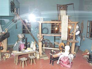 The shop window depicts a miniature cottage with dolls spinning yarn and weaving fabric on a loom