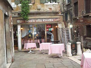 The sign on the awning reads, 'Vini Sceli alla Rivella,' there are tables with pink cloths set out in the narrow street, stacks of chairs waiting to be deployed, a bit of green descending from a potted plant in the window above, and a more formal dining area within the small restaurant