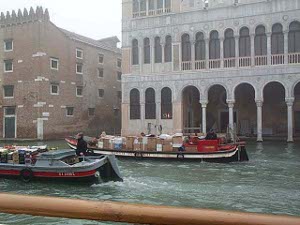 Two motorized delivery barges cruise down a Venetian canal.  The rail of the vaporetto can be seen in the foreground, and famous old Venetian buildings in the background.