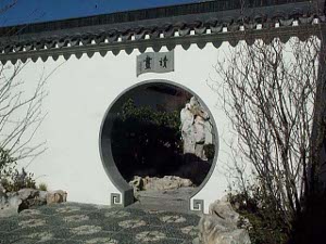 The round door has a gray outline in a white wall, with decorative gray fret-like desings near the ground and Chinese characters above the door; the floor consists of floral designs in stone