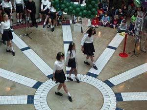 Young girls in white blouses and green tartan skirts, with white socks and black dancing shoes, performing on a tiled dance floor in Lonsdale Quay