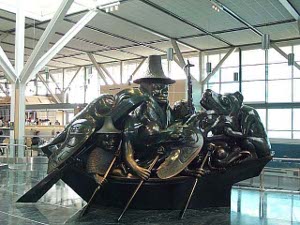 The stylized crowded green canoe is being paddled across the floor of the airport room; the character at the top has a large unforgettable hat, like a truncated inverted funnel