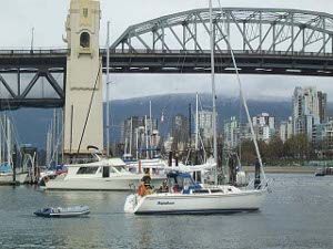 A motor sailboat is putting out for a day sail, towing a tiny dinghy, other boats are tied up at the marina, and the tall bridge towers above