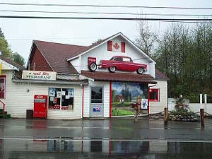 A white building with red trim and an old car mounted on the second floor and the large mural on the wall facing the street, along with old gas pumps