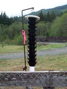 On the other side of a fence, a white cylinder comes up from the ground, then twelve cones, point down in a nest then a white topwith a black iron pole (to which a pink ribbon is fastened