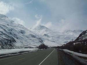 The road surface is wet from a light snow, there are wide shoulders, and tufts of clouds cling to the slopes of the snow covered mountains surrounding the highway