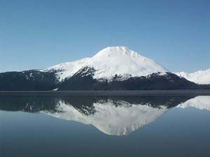 The mountain is dark below and white above with snow; the lake is blue and glassy smooth so the mountain is perfectly reflected in the lake.