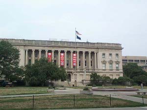 With a tall flagpole in front, and some trees and shrubs around, the Wisconsin Historical Society building is a multi-story tan stone building with a large front gallery set off with a dozen pillars.