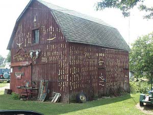 A faded dark red barn with a green shingle roof is decorated with thousands of wrenches hungfrom the walls, mostly vertical