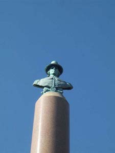 Against a blue sky, the copper green bust of James Avery rests high atop a polished tan stone pillar