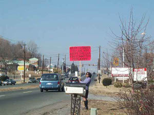 By the side of a divided city road, wearing sunglasses, a purple sweater, black skirt and boots, a dark-haired woman holds a large bright red placard (see text in story).