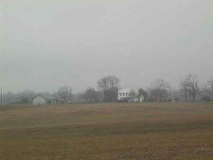 On a gray autumn day, the open field in the foreground has an orange hue, the trees are bare of leaves, and 500 yards away from the photographer is a two-story white farm house, still standing since the Civil War