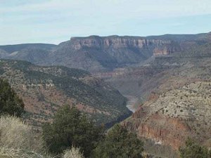 The canyon stretches wide, with cliffs near the top, ocher hillsides dotted with green shrubs and trees, and the narrow river at the bottom of the canyon.