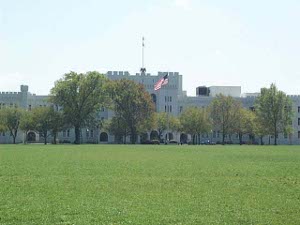Across a broad expanse of green drill field, the white building with arched doorways and crenellated roof flies the American flag.