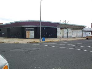 A flat-roofed corner building, painted black and boarded up, sits on a deserted corner of a potholed street in Asbury Park