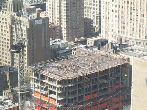 A crane rises high above, while men raise the floors of a skyscraper.  The lower floors are lined with orange plastic to prevent parts from falling to the ground