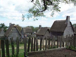 A rough stockade of wooden planks protects yards and gardens, while solid wooden houses with thatched roofs are set side by side with a dirt road down the middle