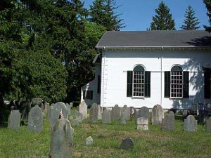 A white wooden building with a gray roof, the back of the Kingston Church is seen, surrounded by evergreens and a small graveyard.