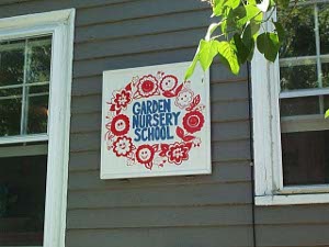 Against a gray clapboard wall, between two white windows, is a white sign with a wreath of bright red flowers surrounding the blue letters 'GARDEN NURSERY SCHOOL'