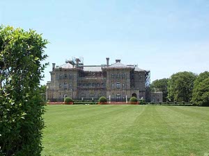 Covered in scaffolding, the brown stone 3-story mansion is seen across a wide and deep expanse of lawn.