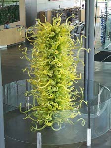 A delicate yellow green glass flower, about four feet high and two feet in diameter, has hundreds of tapering glass tendrils twisting outward from a central core.