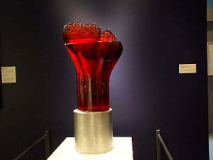 A large abstract red glass flower sits on a polished brass cylindrical base; about two feet high, the light makes shadowy patterns through the dark red glass.