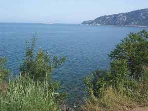 With lakeside grasses in the foreground, and a rocky hillside sloping down from right to left, the blue waters of Lake Superior show small ripples